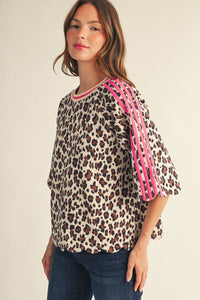 A woman wearing a leopard print top with bright pink shoulder straps and blue jeans, photographed against a plain light beige studio background.