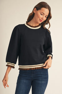 A woman wearing a black long-sleeve knit sweater with cream and brown striped ruffle trim at the crew neck, cuffs, and hem, paired with blue jeans against a light beige studio background.