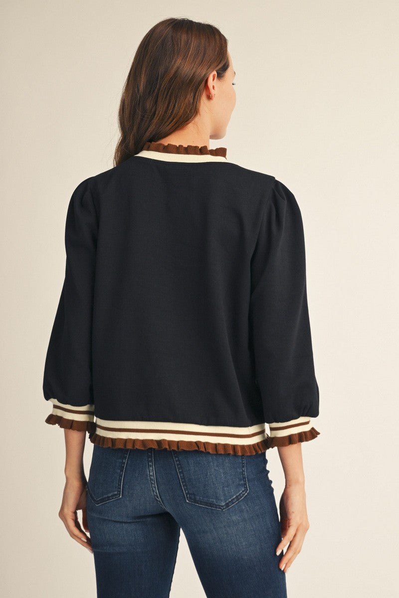 A woman shown from the back wearing a black cropped jacket with brown and cream striped ruffle trim at the collar, cuffs, and hem, paired with blue jeans against a light beige studio background.