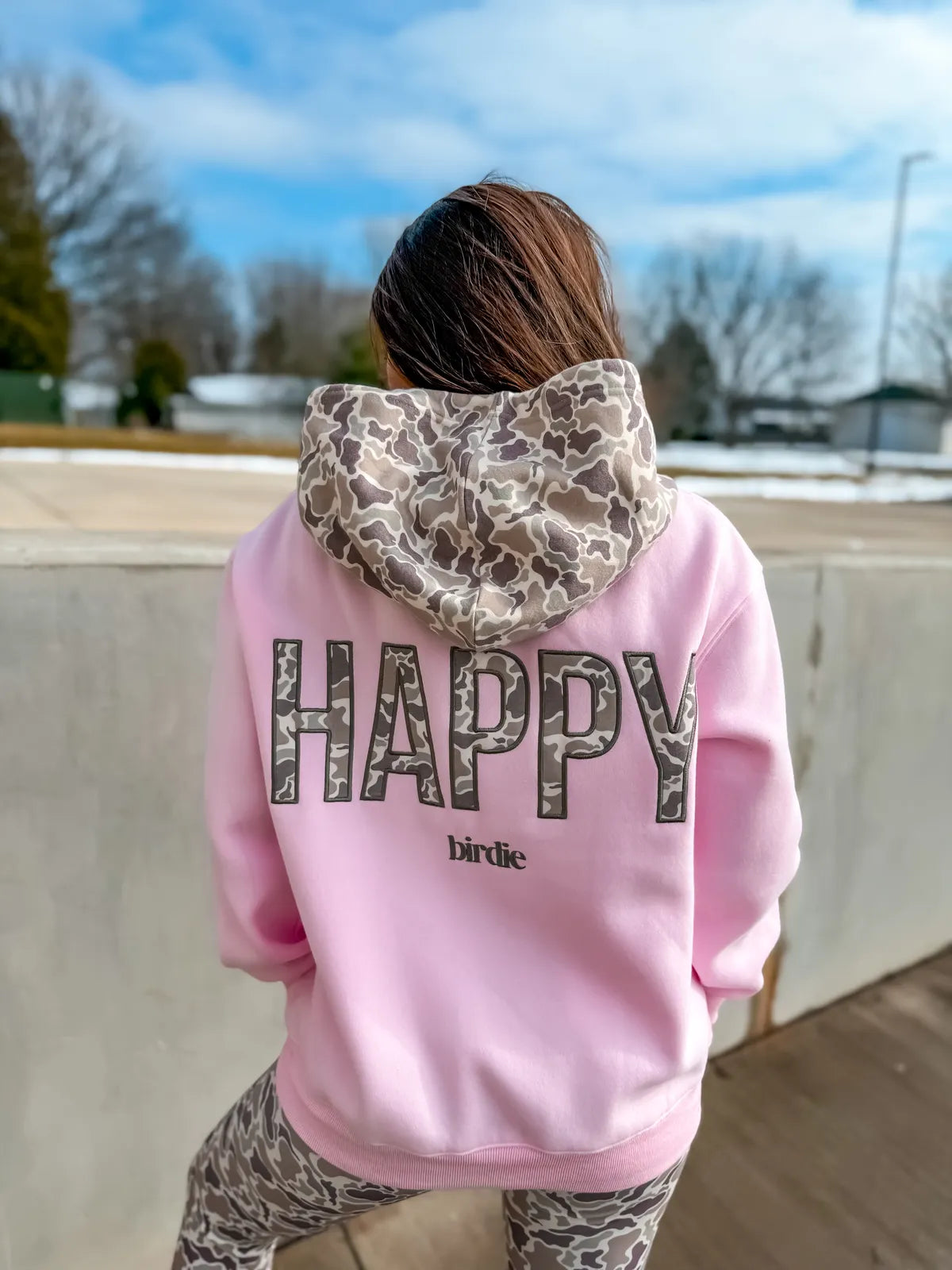 A person viewed from behind wearing a light pink hoodie with a large 'HAPPY' graphic on the back in a gray camouflage pattern, and a matching camouflage hood lining. The brand name 'birdie' is printed below the graphic.