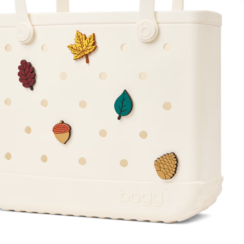 An off-white Bogg reusable tote bag with decorative autumn-themed charms including a yellow maple leaf, dark red leaf, teal leaf, acorn, and pinecone, shown against a white background.