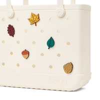 An off-white Bogg reusable tote bag with decorative autumn-themed charms including a yellow maple leaf, dark red leaf, teal leaf, acorn, and pinecone, shown against a white background.