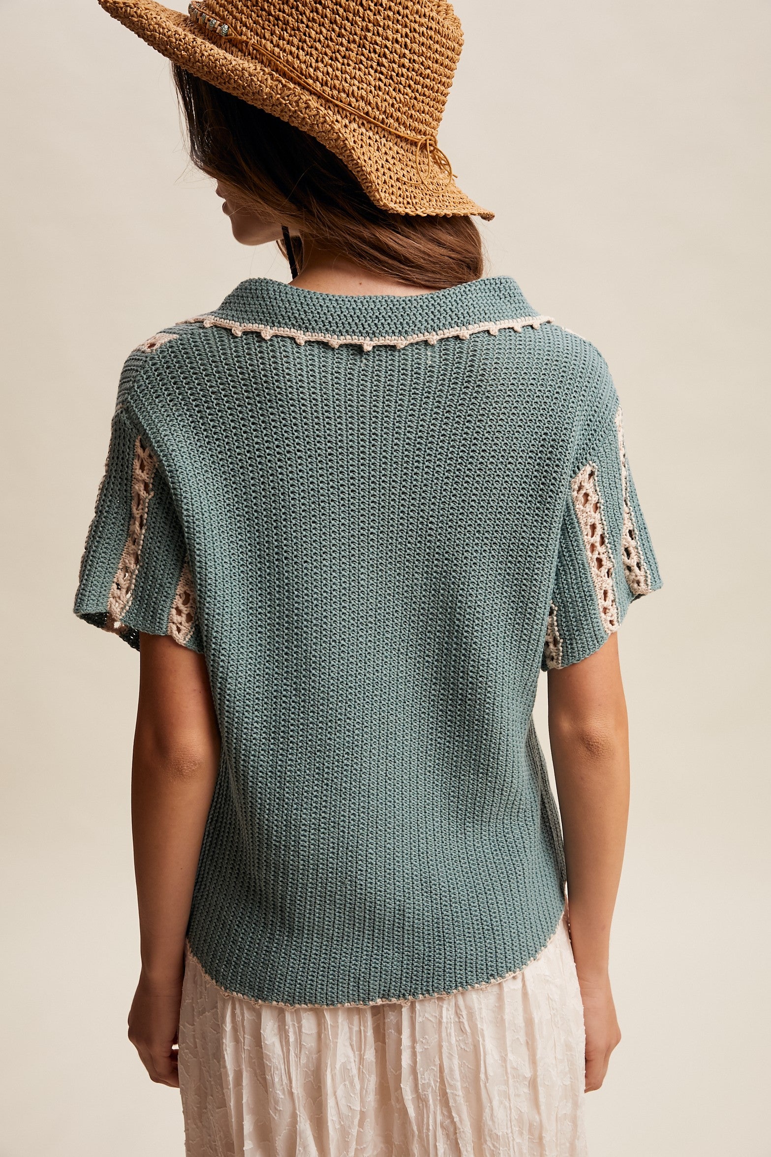 Woman wearing a teal knitted top with lace details and a straw hat on a neutral background