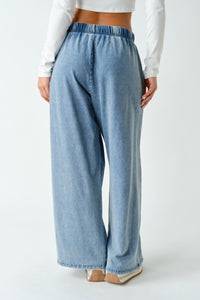A person shown from the back wearing light blue wide-leg denim pants with an elastic waistband, paired with a white long-sleeve crop top and beige heels, against a plain light gray studio background.