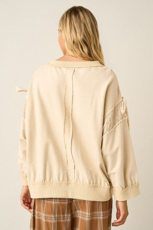A woman shown from the back wearing a cream-colored knit sweater with lace sleeve details and a tie at the collar, paired with brown plaid pants, against a plain light beige background.