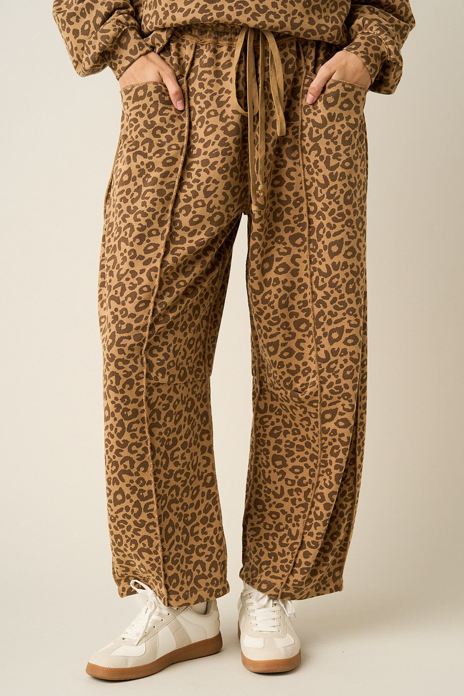 A person wearing loose, wide-leg leopard-print pants with a drawstring waist, shown from the waist down against a plain light beige studio background. The pants have a tan and brown animal pattern with dark brown spots.