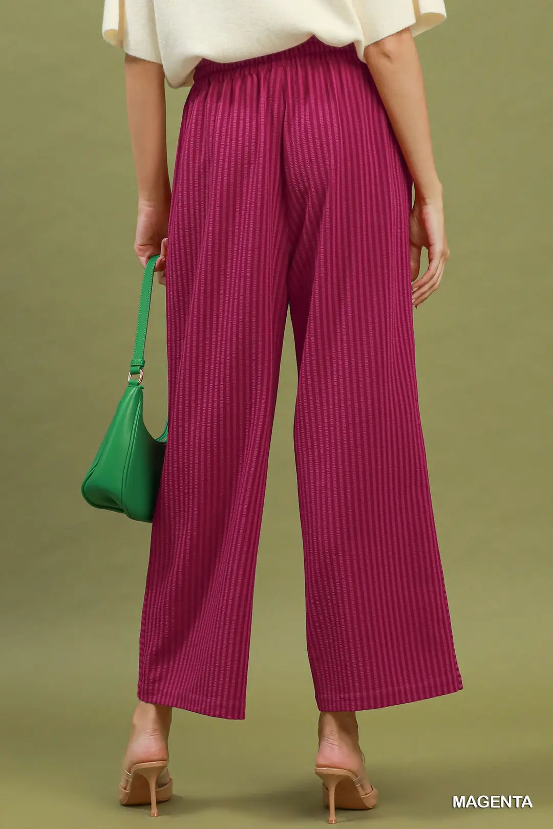 A person shown from the back wearing bright magenta wide-leg pleated pants, a cream short-sleeve top, and nude heels, standing against an olive green studio background and holding a green handbag. The word MAGENTA appears in the lower right corner.