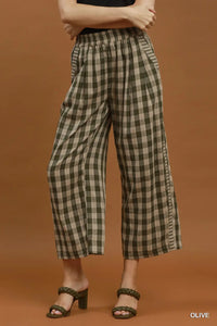 A model wearing olive green and beige checkered cropped wide-leg pants with a black top and dark green heeled sandals, set against a solid warm brown studio background.