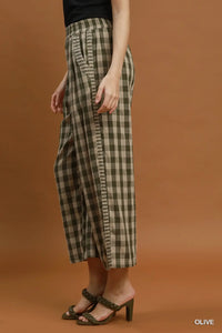 A model wearing olive green and beige checkered cropped wide-leg pants with side pleats, paired with dark green heeled sandals, against a solid warm brown studio background.