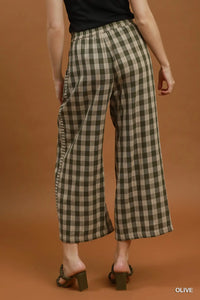 A model from the back wearing olive green and beige plaid wide-leg cropped pants, paired with a black top and dark green block-heel slide sandals, against a solid warm brown studio background.