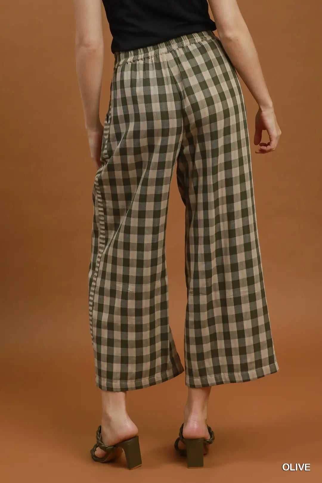 A model from the back wearing olive green and beige plaid wide-leg cropped pants, paired with a black top and dark green block-heel slide sandals, against a solid warm brown studio background.