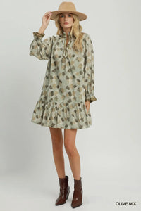 A woman modeling a long-sleeve mini dress with an olive green, beige, and brown abstract pattern, paired with a tan wide-brim hat and brown ankle boots, against a light gray studio background.