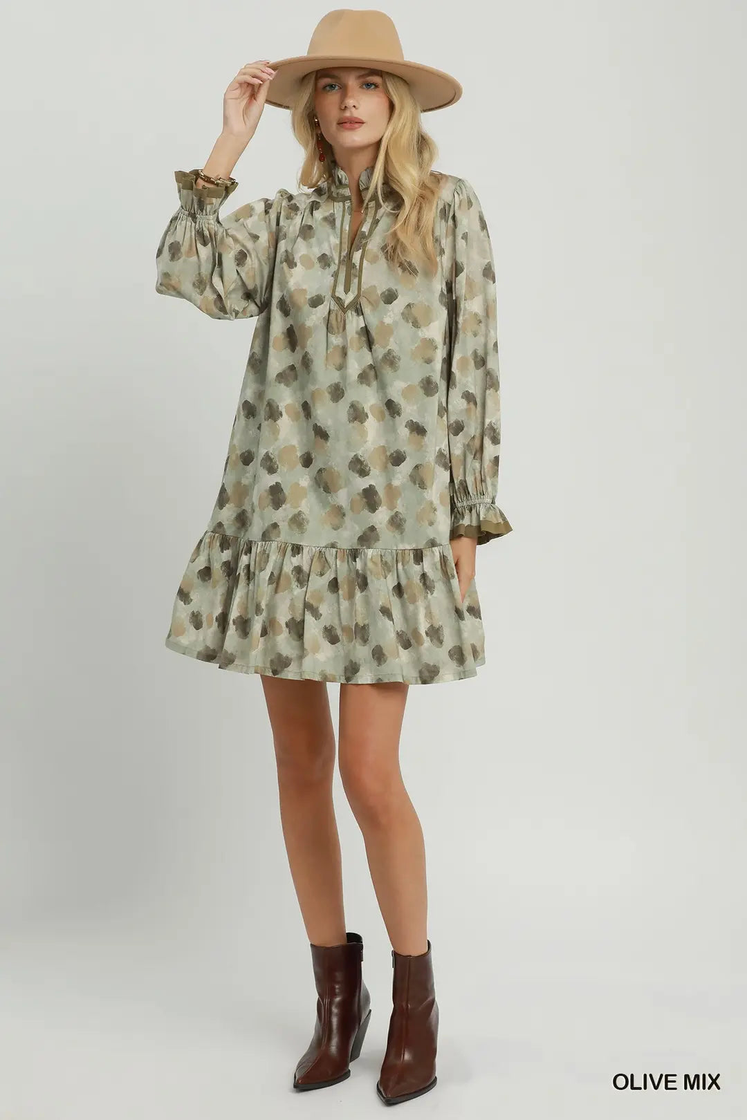 A woman modeling a long-sleeve mini dress with an olive green, beige, and brown abstract pattern, paired with a tan wide-brim hat and brown ankle boots, against a light gray studio background.