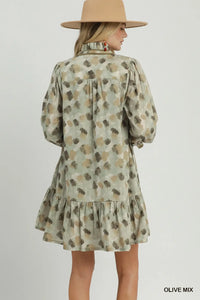 A woman shown from the back wearing a short olive green dress with a brown and beige abstract spotted pattern, long puff sleeves, and a ruffled hem. She is also wearing a wide-brimmed tan hat.