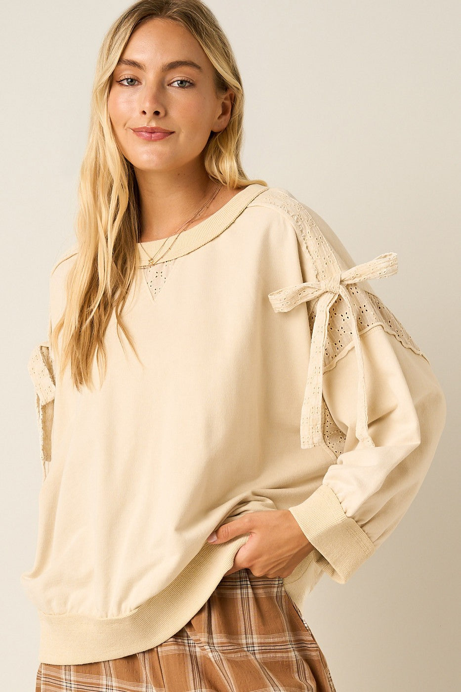 A woman wearing a cream-colored long-sleeve top with lace-up bow details on the sleeves and a brown plaid skirt, photographed against a solid light beige background.
