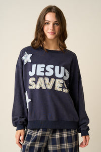 A woman wearing a navy blue crewneck sweatshirt with 'JESUS SAVES' printed in white and glittery gold letters, paired with navy plaid pants, against a solid beige background.