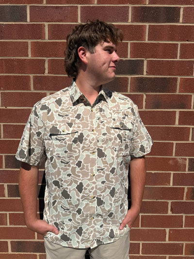A man wearing a short-sleeve button-up shirt with a beige, brown, and gray camouflage pattern, standing against a red brick wall.