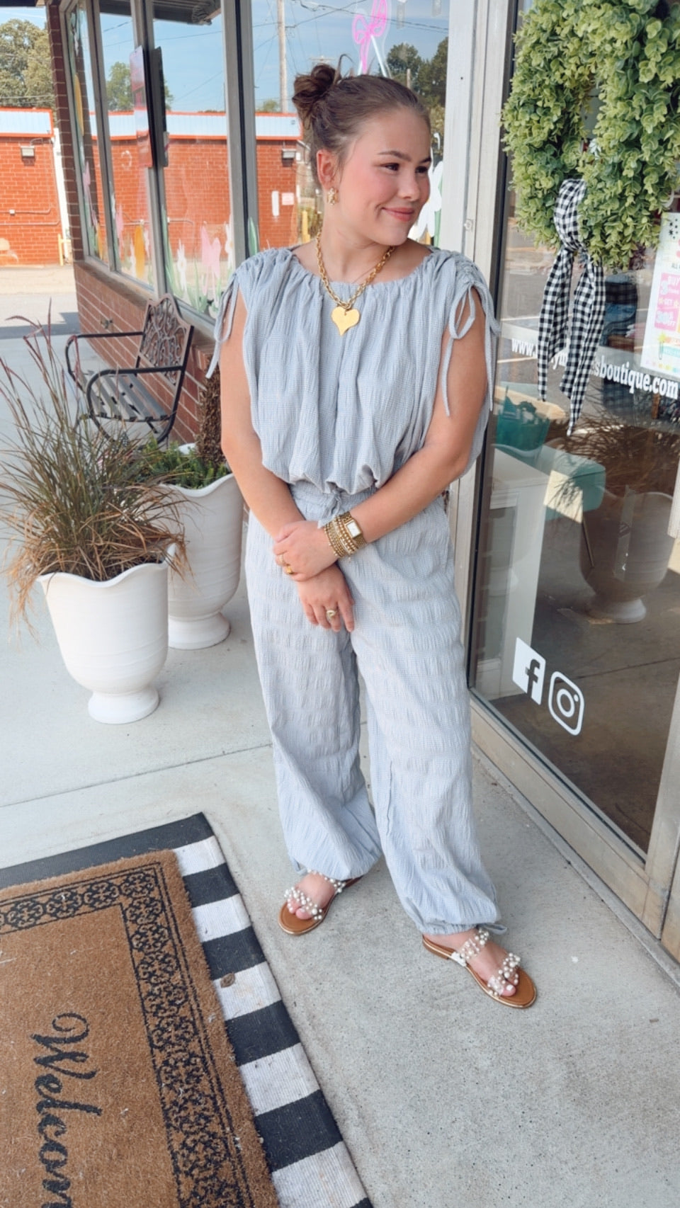 A woman wearing a light blue-gray sleeveless jumpsuit with wide-leg pants and brown sandals, standing on a storefront porch.