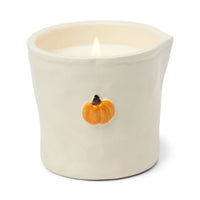 A lit cream-colored ceramic candle holder with a small orange pumpkin decoration on the front, set against a plain white background.