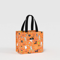 An orange tote bag with a black handle and a Halloween-themed print featuring cartoon skeletons, ghosts, pumpkins, and cats, shown on a light gray background.