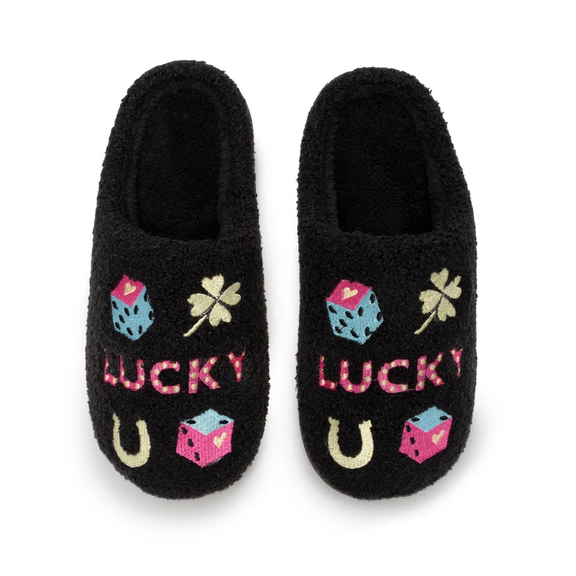 A pair of black fuzzy slip-on slippers with St. Patrick's Day themed embroidery, including pink and blue dice, shamrocks, horseshoes, and the word 'LUCKY' in pink, shown on a white background.