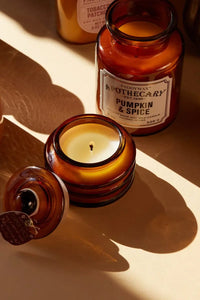 Amber glass jar candle shown open with its lid beside it on a warm beige surface, with a second matching candle in the background. The front candle label reads 'Paddywax Apothecary Candle Pumpkin & Spice'.