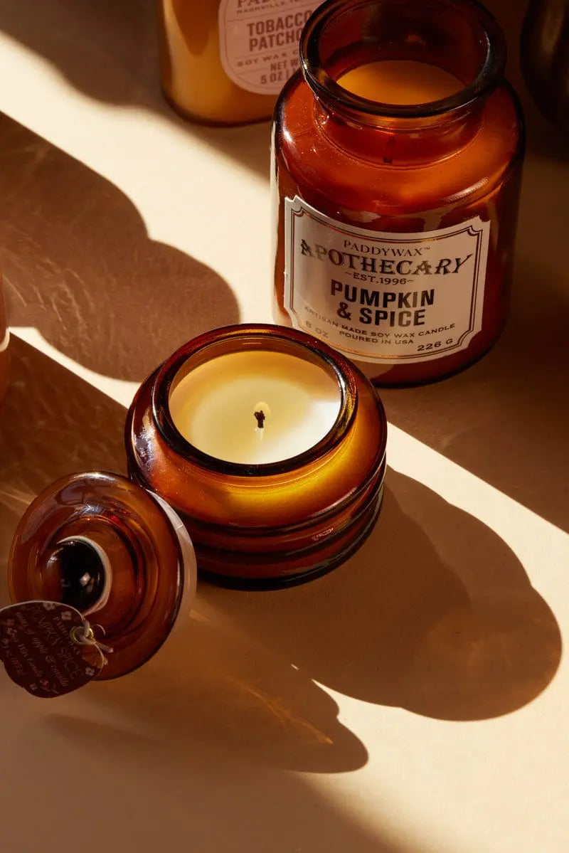 Amber glass jar candle shown open with its lid beside it on a warm beige surface, with a second matching candle in the background. The front candle label reads 'Paddywax Apothecary Candle Pumpkin & Spice'.