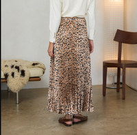 A woman wearing a pleated midi skirt with a beige, brown, and black leopard print pattern, paired with a white long-sleeve top and brown flat sandals, standing in a modern room.