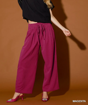 A model wearing magenta wide-leg pinstriped pants with a drawstring waist, paired with a black top and pink heels, standing against a solid brown background.
