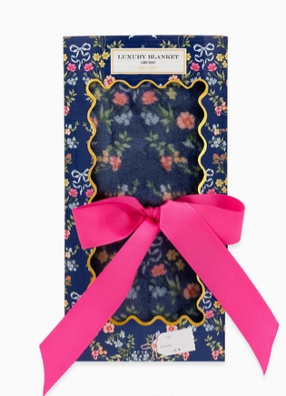 A packaged floral blanket with a bright pink ribbon bow. The product is shown in its retail packaging, featuring a navy blue floral patterned blanket with a gold scalloped border, set against a clean white background.