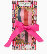 A packaged plush blanket gift set with a bright pink ribbon bow. The box is decorated with pink and white stripes, cartoon dog illustrations, and a gold-trimmed window showing a folded blanket with pink, orange, and blue stripes.