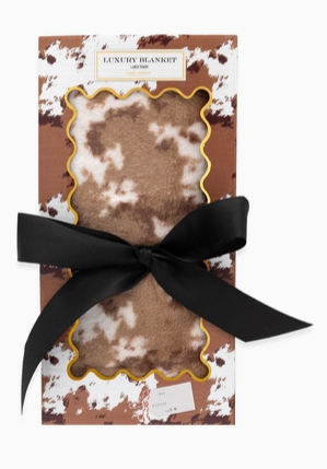 A luxury blanket gift set featuring a brown and white cow-print fleece blanket with a scalloped gold border, presented in a brown and white cow-print box with a black ribbon bow.