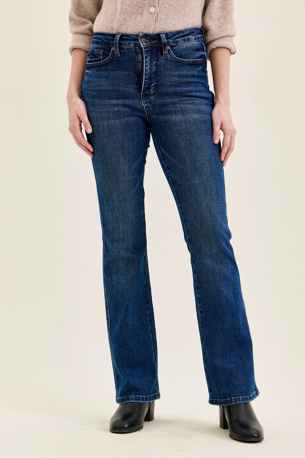 A person wearing dark blue straight-leg denim jeans with a beige cardigan and black ankle boots, shown from the waist down against a plain light beige studio background.