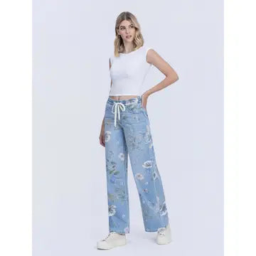 A woman modeling light blue wide-leg jeans with a floral print and white drawstring waist, paired with a white sleeveless crop top and white sneakers, against a light gray studio background.