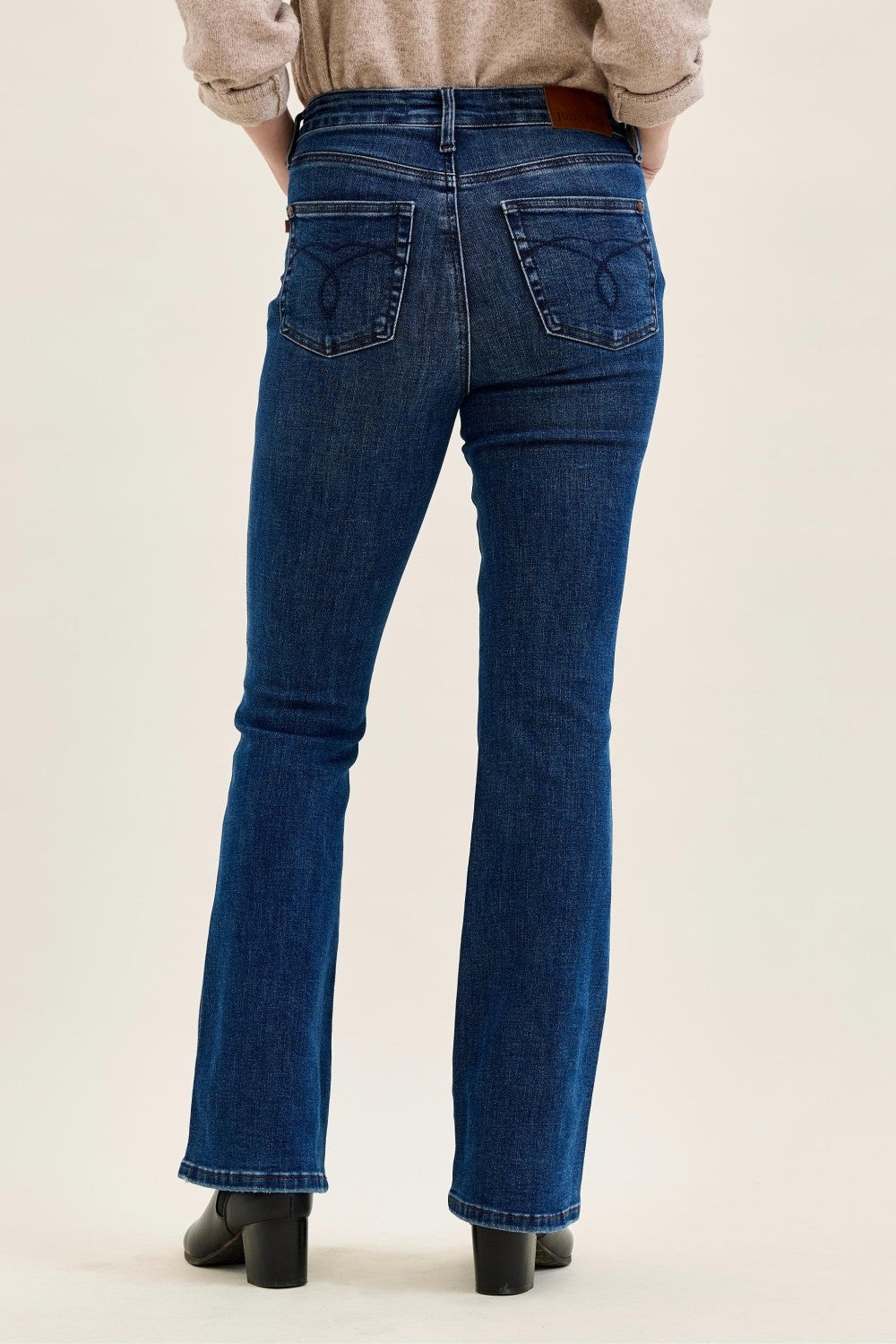 Back view of a person wearing dark blue denim flare jeans with a brown leather waistband and black block-heel boots, against a plain light beige studio background.