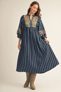A woman modeling a navy blue maxi dress with vertical white stripes, a colorful embroidered yoke, and patterned sleeve cuffs, paired with brown boots against a light beige studio background.
