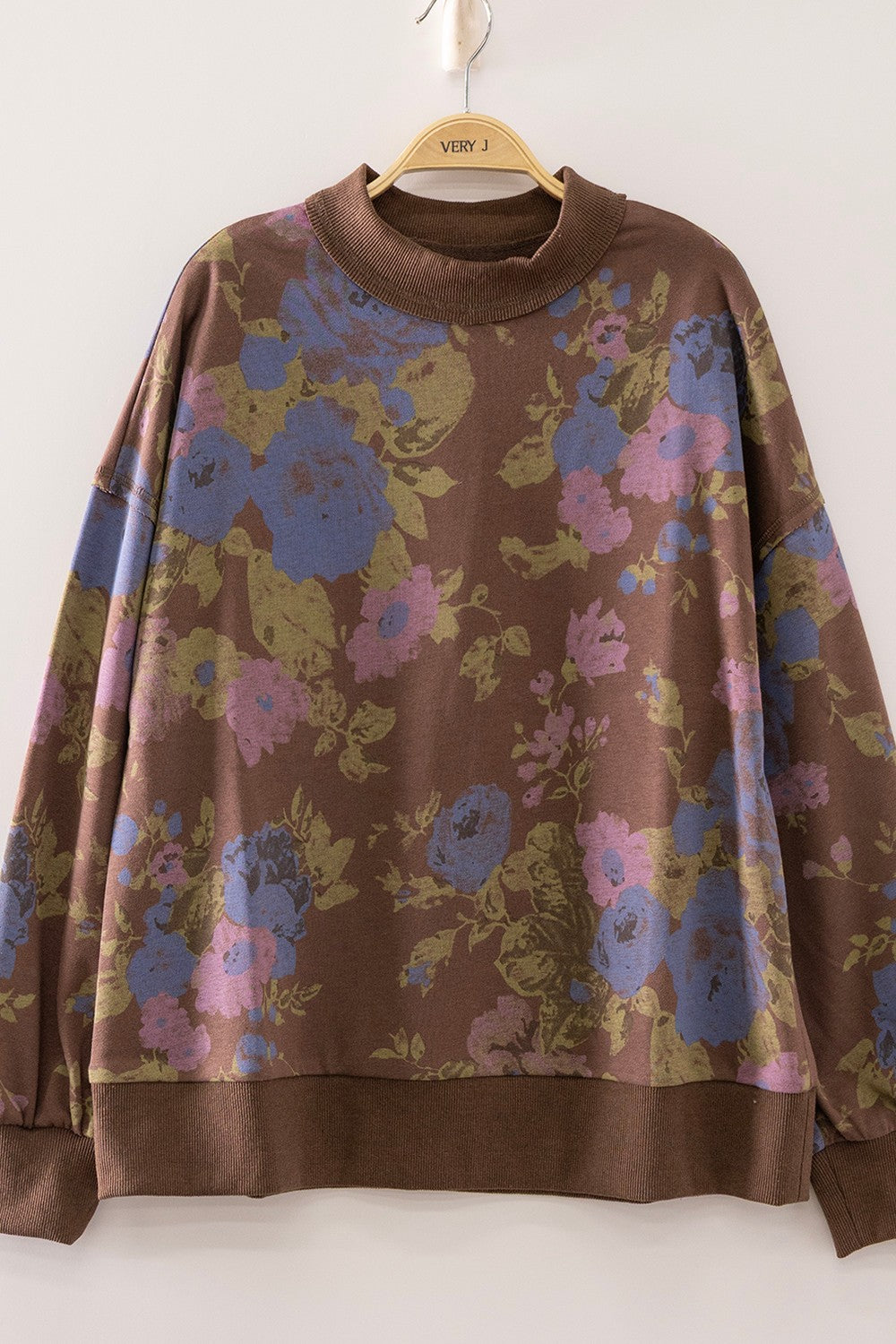 A brown long-sleeve sweatshirt with a large floral pattern in blue, pink, and olive green, hanging on a wooden hanger against a plain white wall.