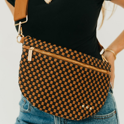 A close-up of a person wearing a woven brown and black checkered crossbody bag with gold-tone hardware, a black top, and blue jeans against a plain white background.