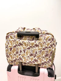 A beige, brown, and white camouflage patterned soft-sided bag with black handles, stacked on top of a pink hard-shell suitcase against a plain off-white wall.