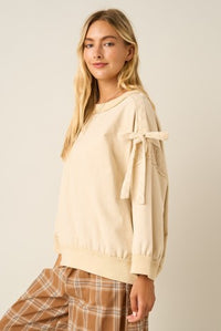 A woman wearing a cream-colored knit sweater with lace-up bow details on the sleeves, paired with brown and beige plaid pants, set against a plain light beige background.